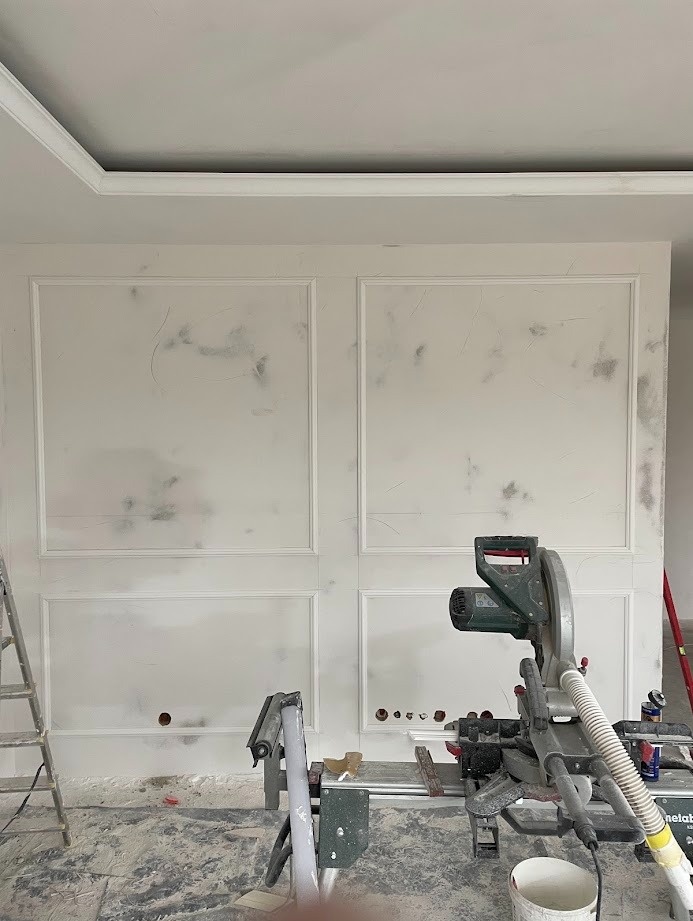 Ściana z dekoracyjnymi panelami w stylu klasycznym, widoczna piła ukośnica i drabina, sugerujące prace wykończeniowe w trakcie realizacji.