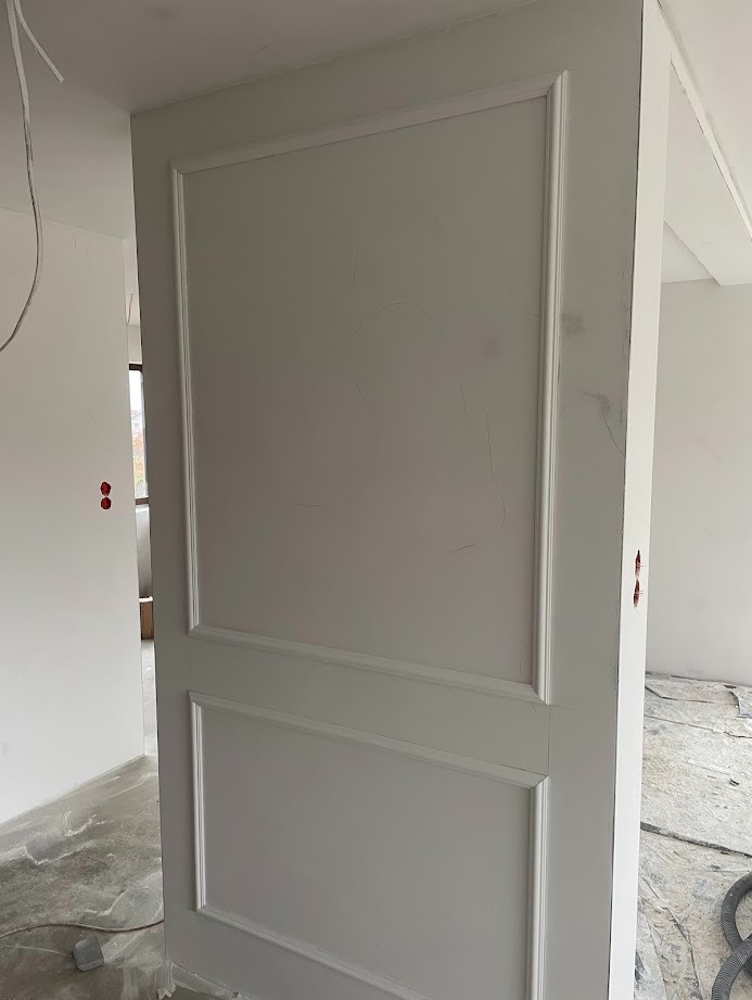 Biała ściana z dekoracyjnymi panelami w stylu klasycznym, widoczne ślady szpachlowania, surowy beton na podłodze, wnętrze w trakcie wykończenia.