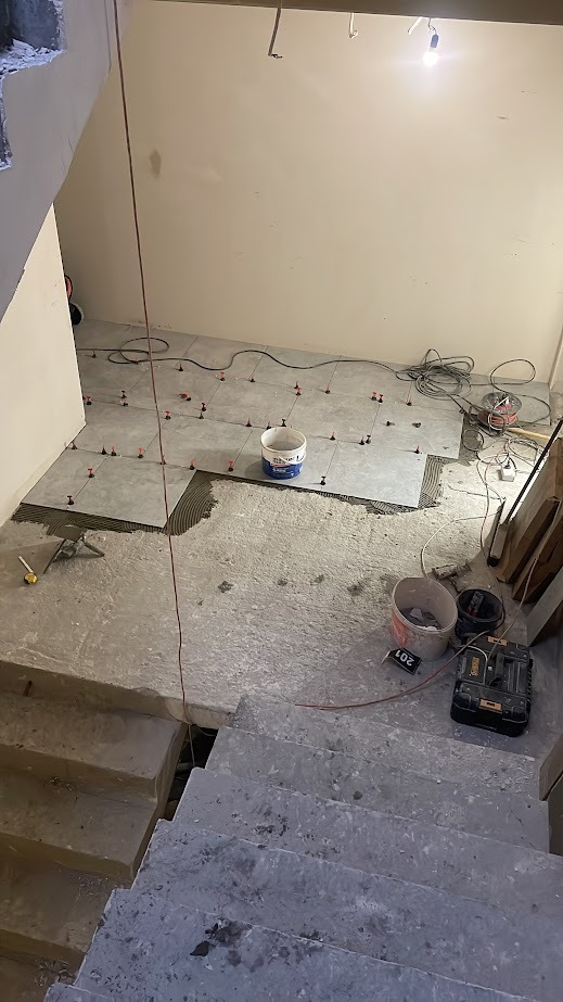 Układanie szarych płytek podłogowych w pomieszczeniu, widoczne narzędzia i akcesoria glazurnicze, etap prac wykończeniowych, surowy stan schodów w tle.