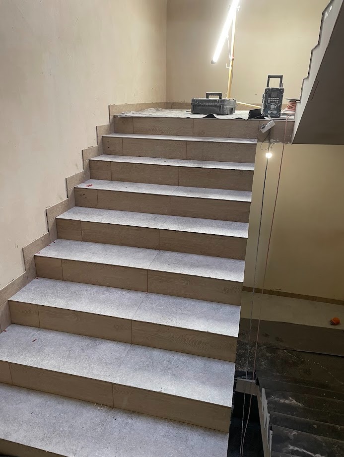 Wykończone schody z jasnymi płytkami imitującymi drewno, widoczne narzędzia i lampa budowlana, ściany w odcieniach beżu. Częściowo widoczne schody przed wykończeniem.