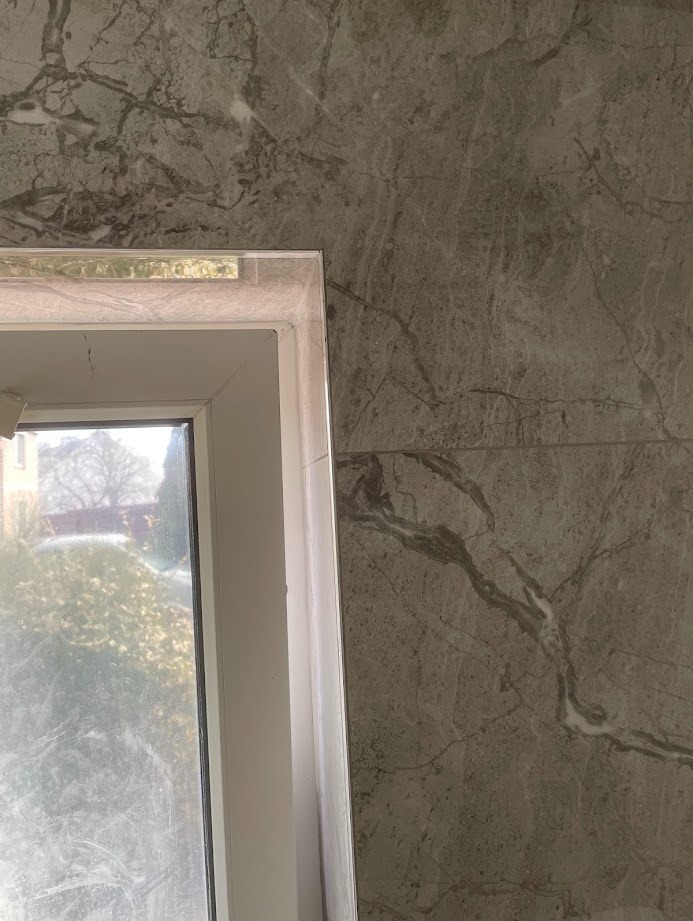 Szara ściana z marmurowym wzorem, obok biała rama okienna. Widok na zewnątrz przez brudną szybę. Minimalistyczny design, neutralna kolorystyka.