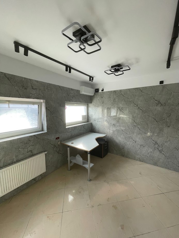 Pokój biurowy z marmurowymi ścianami, biurkiem narożnym i nowoczesnym oświetleniem sufitowym. Widoczne okna i grzejnik. Jasne płytki podłogowe.