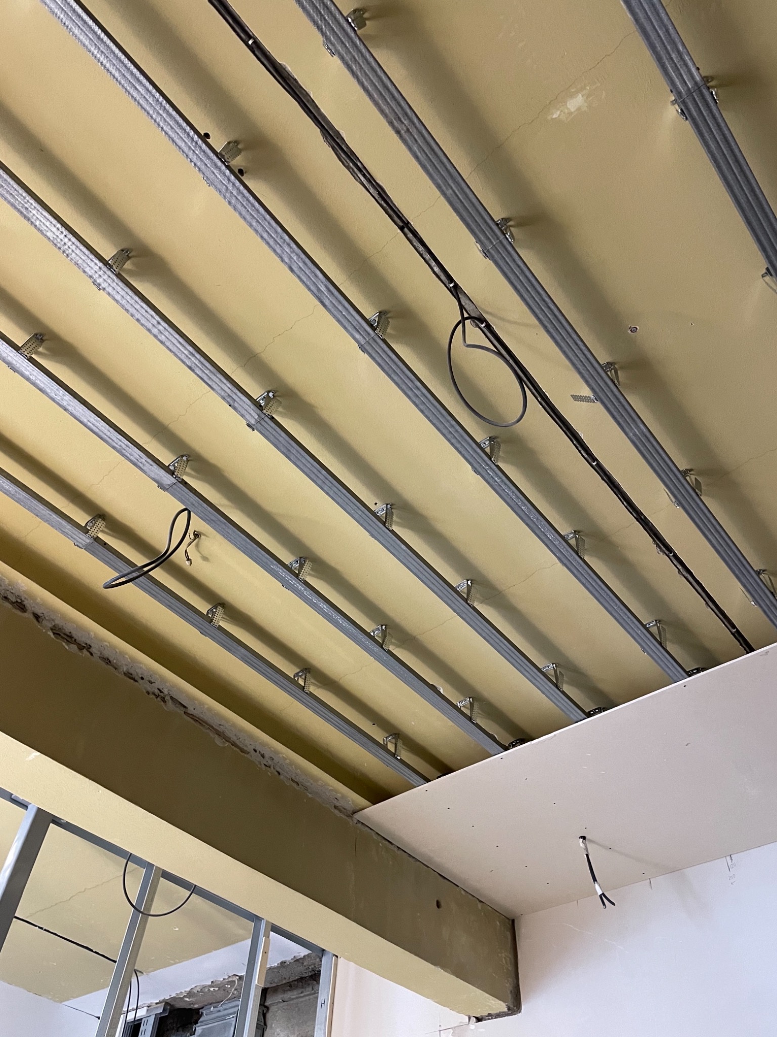 Metalowy stelaż podwieszanego sufitu z płyt gipsowo-kartonowych, widoczne przewody elektryczne i fragment ściany z niedokończonym wykończeniem.