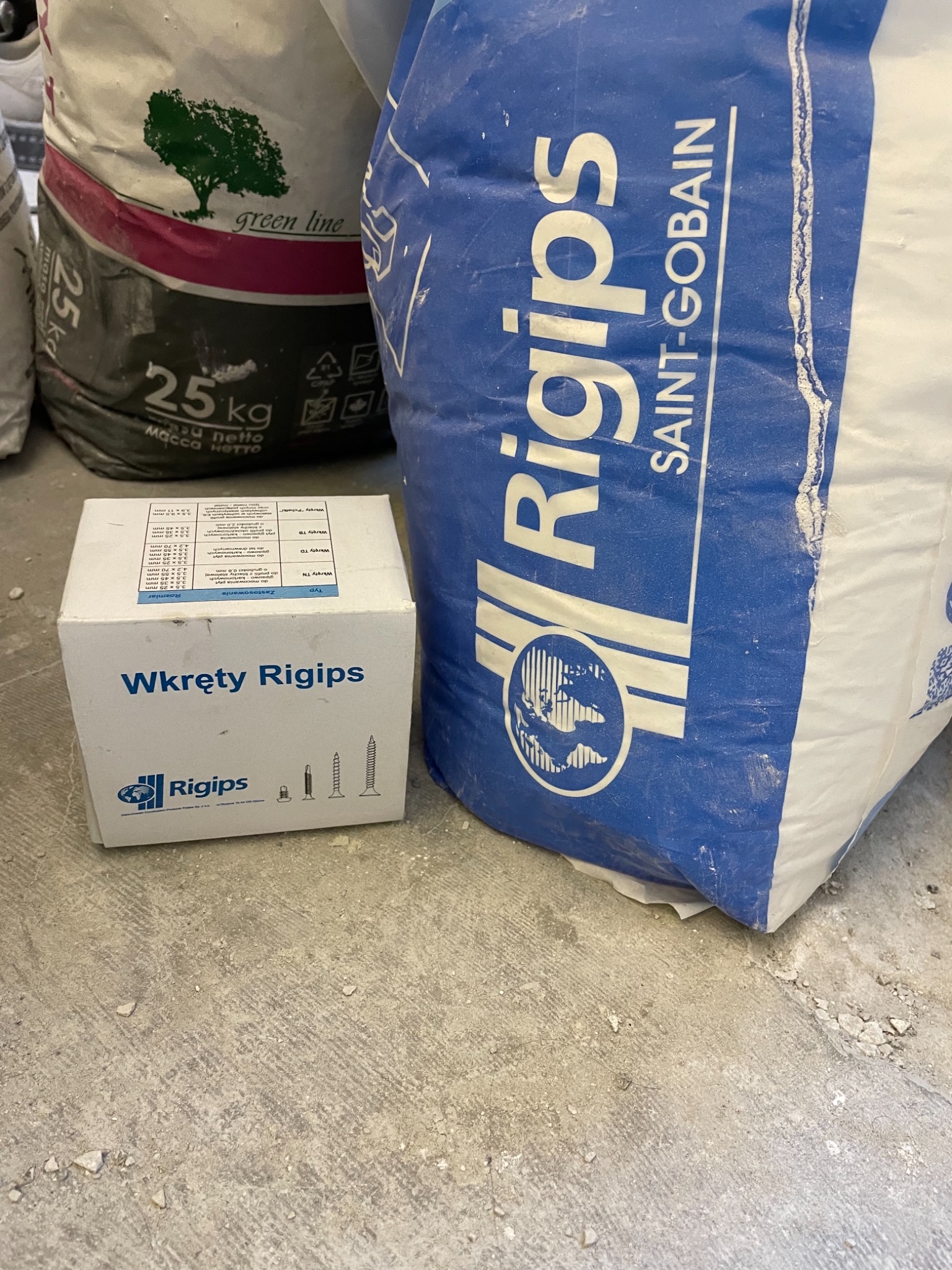 Pudełko wkrętów Rigips oraz dwa worki zaprawy gipsowej Rigips i Green Line leżące na podłodze z resztkami gipsu.