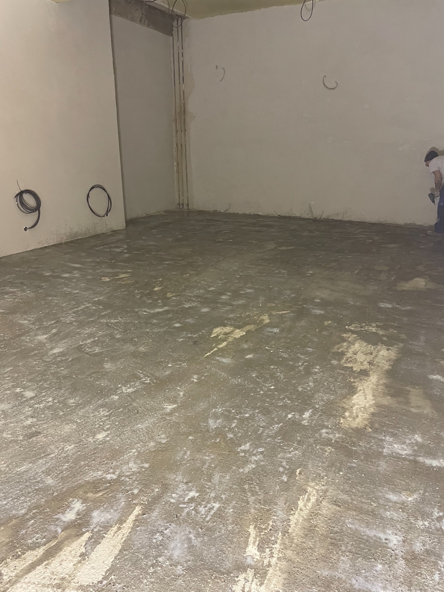 Surowy betonowy podkład podłogowy w trakcie wyrównywania, widoczne ślady białego mleczka cementowego, kable elektryczne zwisające ze ściany, fragment sylwetki pracownika w prawym rogu.