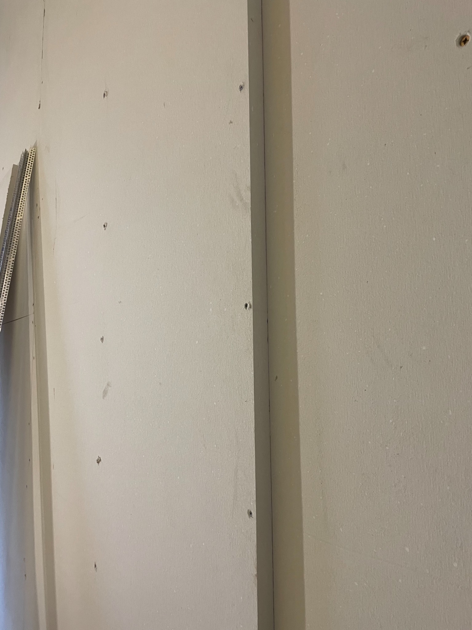 Ściana z płyt gipsowo-kartonowych, widoczne łączenia i wkręty, przygotowana do dalszych prac wykończeniowych, z widocznymi narożnikami aluminiowymi.