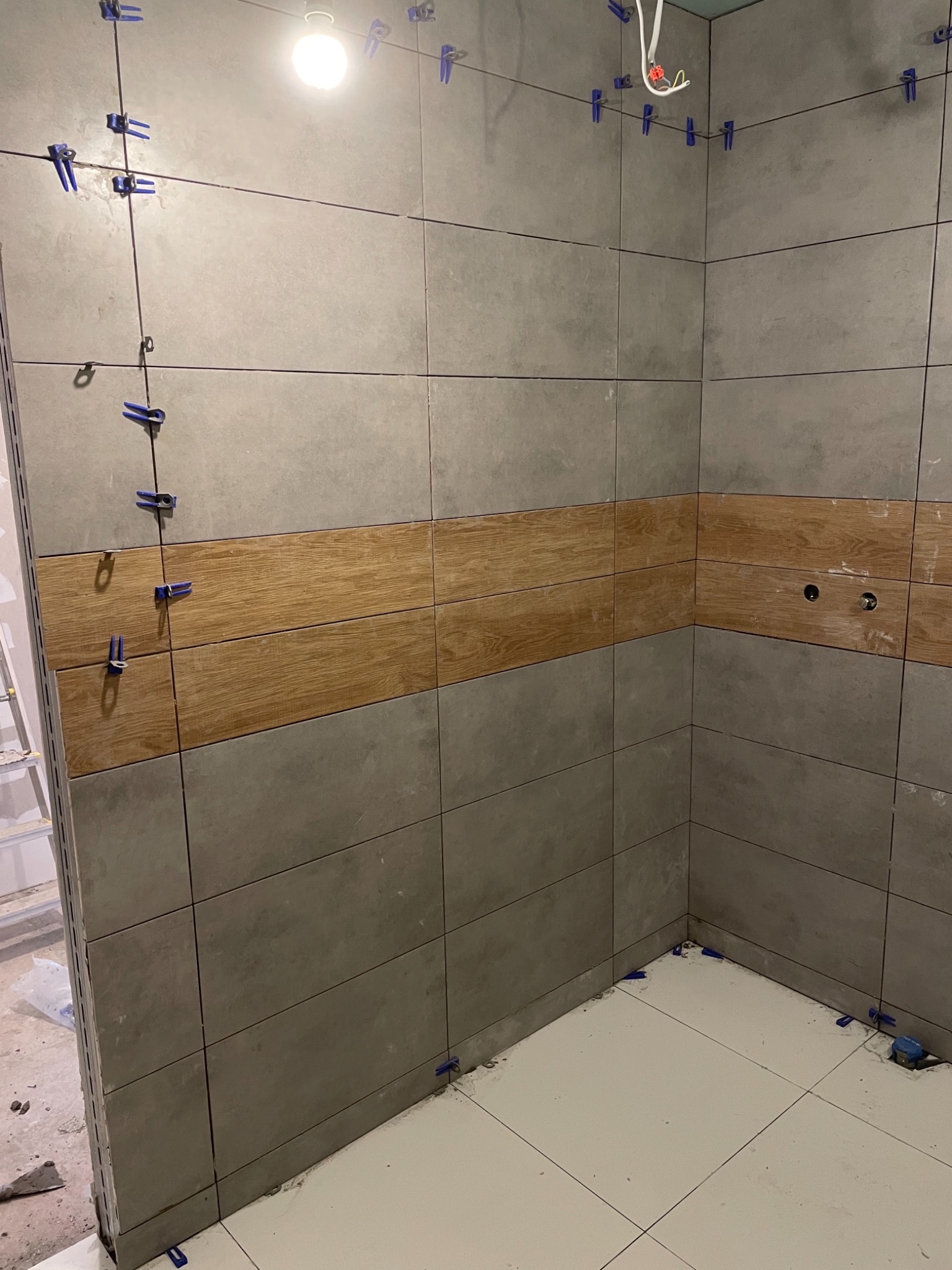 Ściany łazienki w trakcie układania płytek, z szarymi płytkami i poziomym pasem płytek imitujących drewno, użyte klipsy poziomujące, widoczne otwory na armaturę.