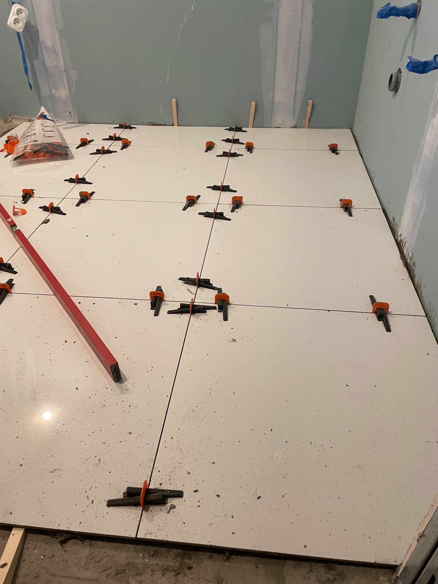 Białe płytki podłogowe w trakcie układania, z systemem poziomowania płytek i poziomicą, widoczne niedokończone ściany w tle.