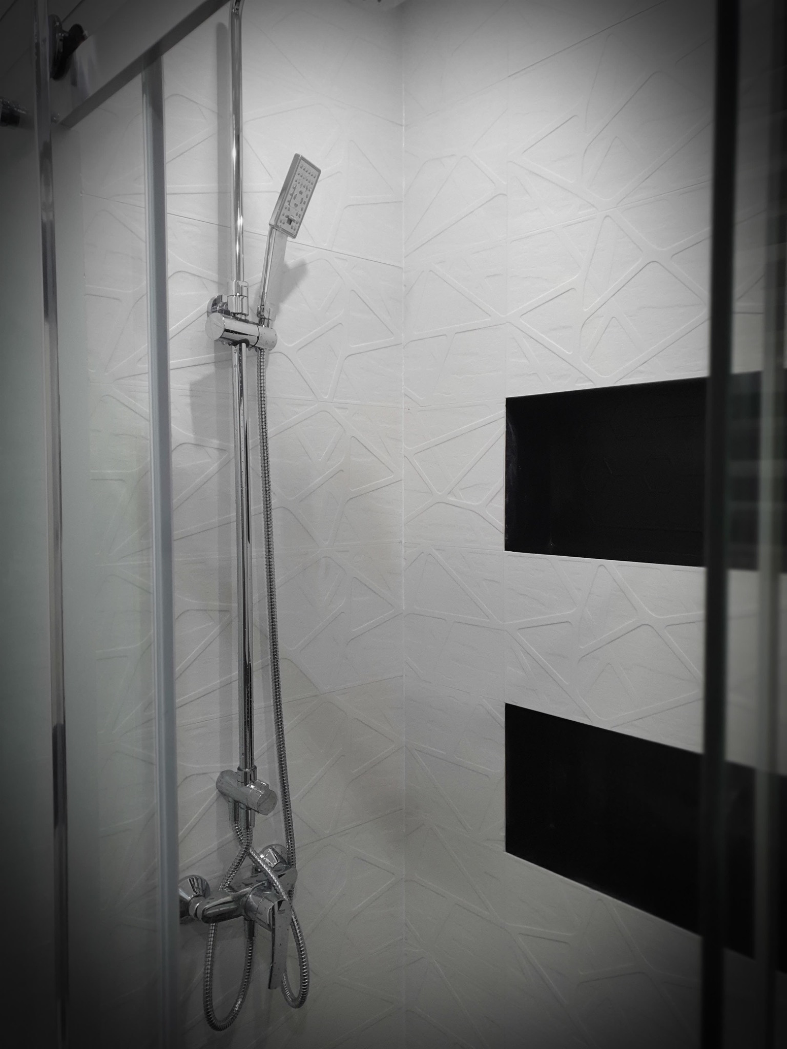Nowoczesna kabina prysznicowa z chromowaną armaturą i biało-czarną mozaiką na ścianie, widoczne geometryczne wzory na płytkach i częściowo przesłonięte szklane drzwi.