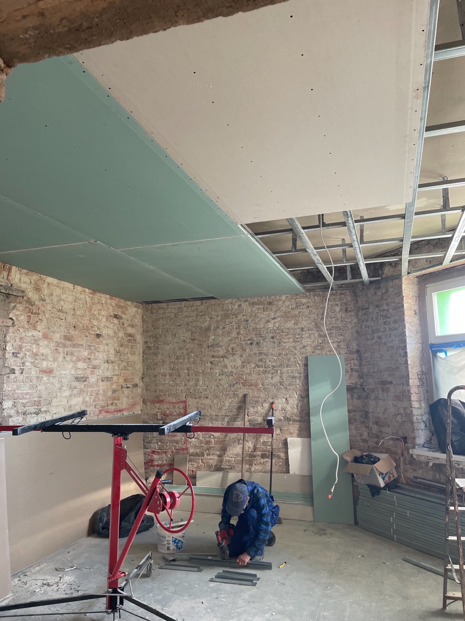 Montaż płyt gipsowo-kartonowych na suficie w pomieszczeniu z surową cegłą, widoczny podnośnik do płyt i pracownik przycinający profile.