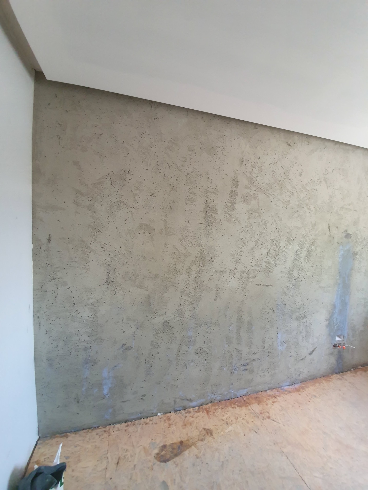 Ściana pokryta szarym tynkiem dekoracyjnym o nieregularnej fakturze, widoczne złącze elektryczne, surowa podłoga z płyt OSB, biały sufit z listwą dekoracyjną.