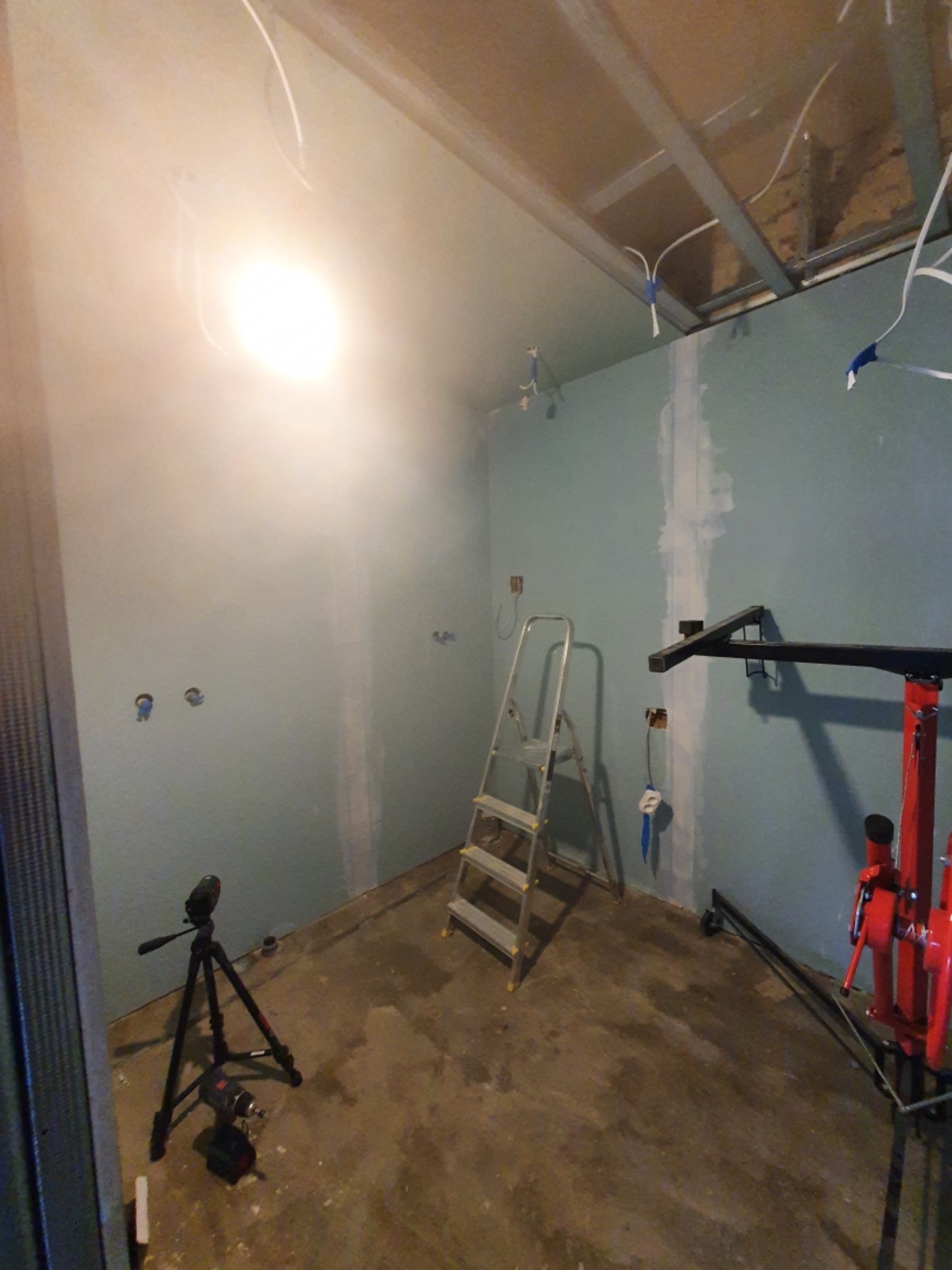 Wnętrze w trakcie remontu: ściany z płyt gipsowo-kartonowych, częściowo zaszpachlowane, widoczne przewody elektryczne, drabina aluminiowa, podnośnik do płyt i poziomica na statywie.