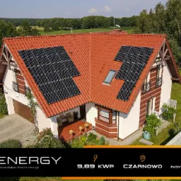 Dom jednorodzinny z czerwoną dachówką, na której zamontowano panele słoneczne w trzech sekcjach, widok z góry, w tle zieleń drzew.