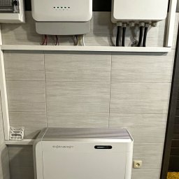 Ozenergy Sp. z o.o. - Biały magazyn energii SiGenergy zamontowany na ścianie z szarych płytek, widoczne kable i elementy instalacji elektrycznej. Domowa instalacja wewnątrz budynku.