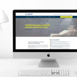 Ekran komputera iMac wyświetlający stronę internetową z nagłówkiem 'Inwestowanie w wiedzę zawsze przynosi zyski', obok biała lampka biurkowa i klawiatura na białym biurku.