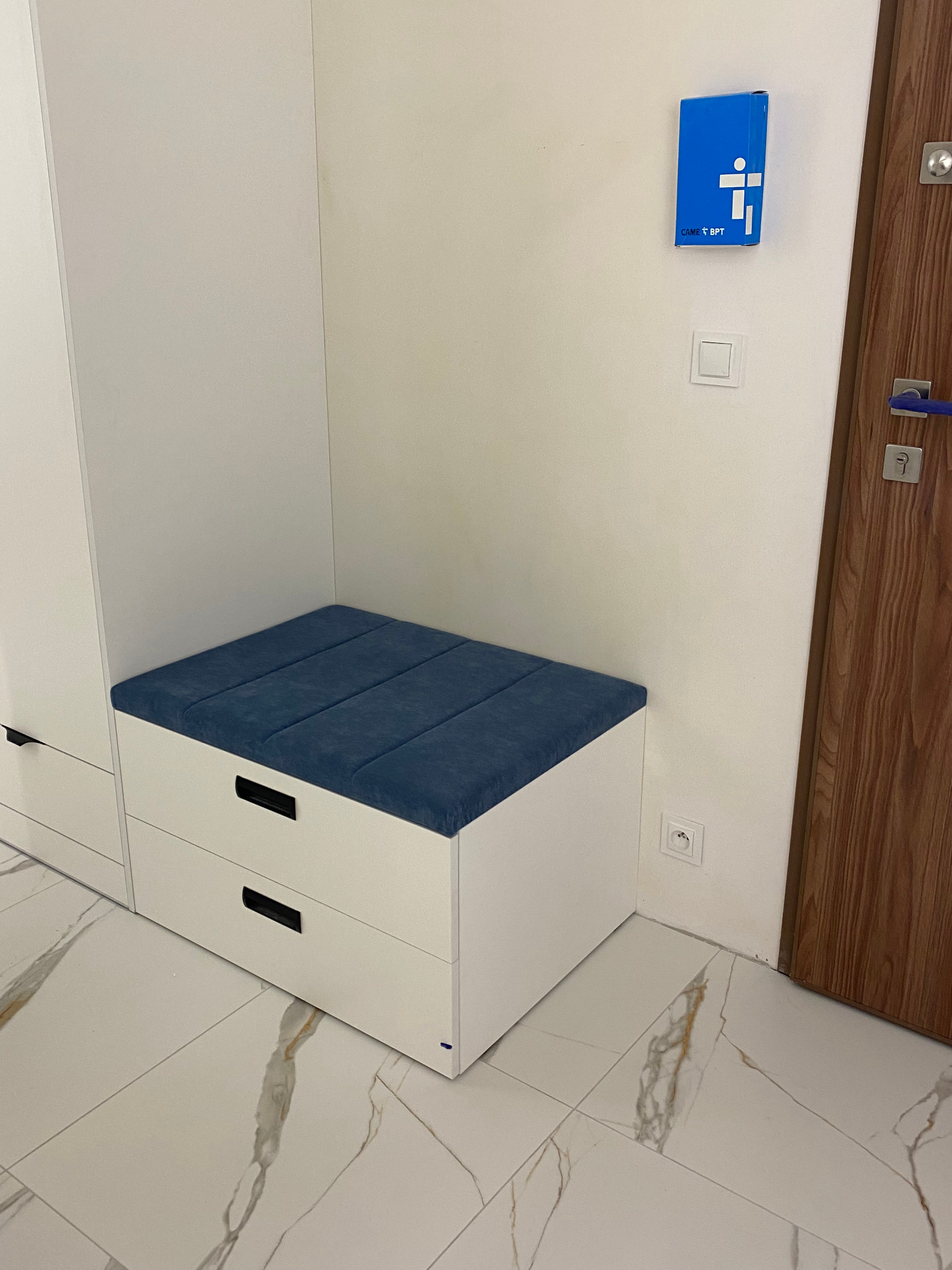 Biała szafka z szufladami i niebieskim siedziskiem w przedpokoju, widoczny fragment szafy, drewniane drzwi, marmurowe płytki podłogowe i niebieskie pudełko na ścianie.