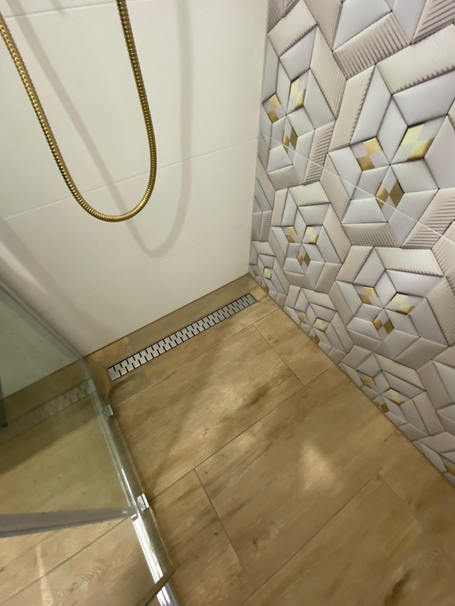 Wykończona łazienka z drewnopodobną podłogą, odpływem liniowym, geometrycznymi płytkami ściennymi z akcentami w kolorze złota oraz złotym wężem prysznicowym.