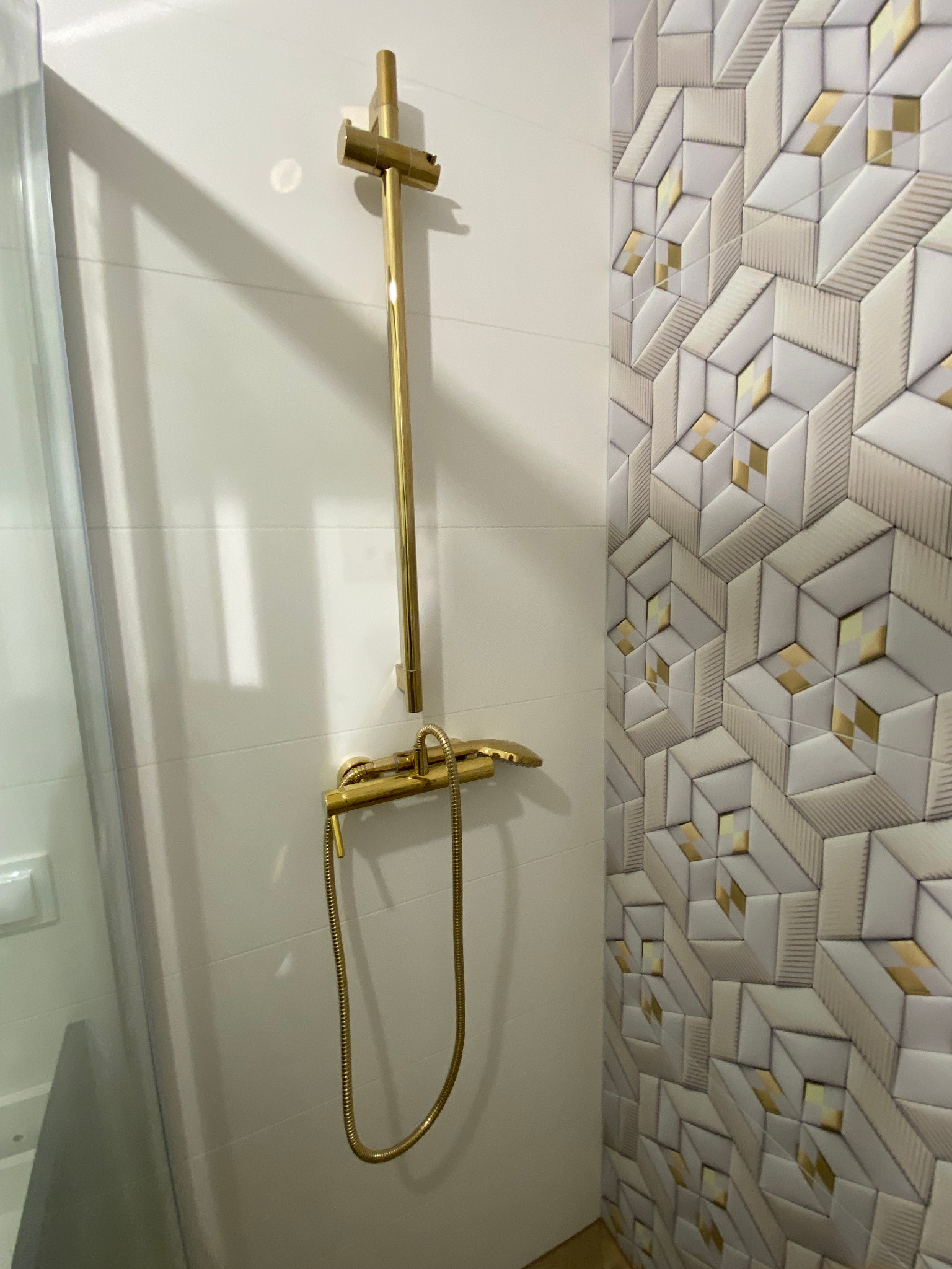 Złota armatura prysznicowa na tle białych płytek z geometrycznym wzorem z elementami w kolorze złota. Widoczna kabina prysznicowa z elementami białej ściany.