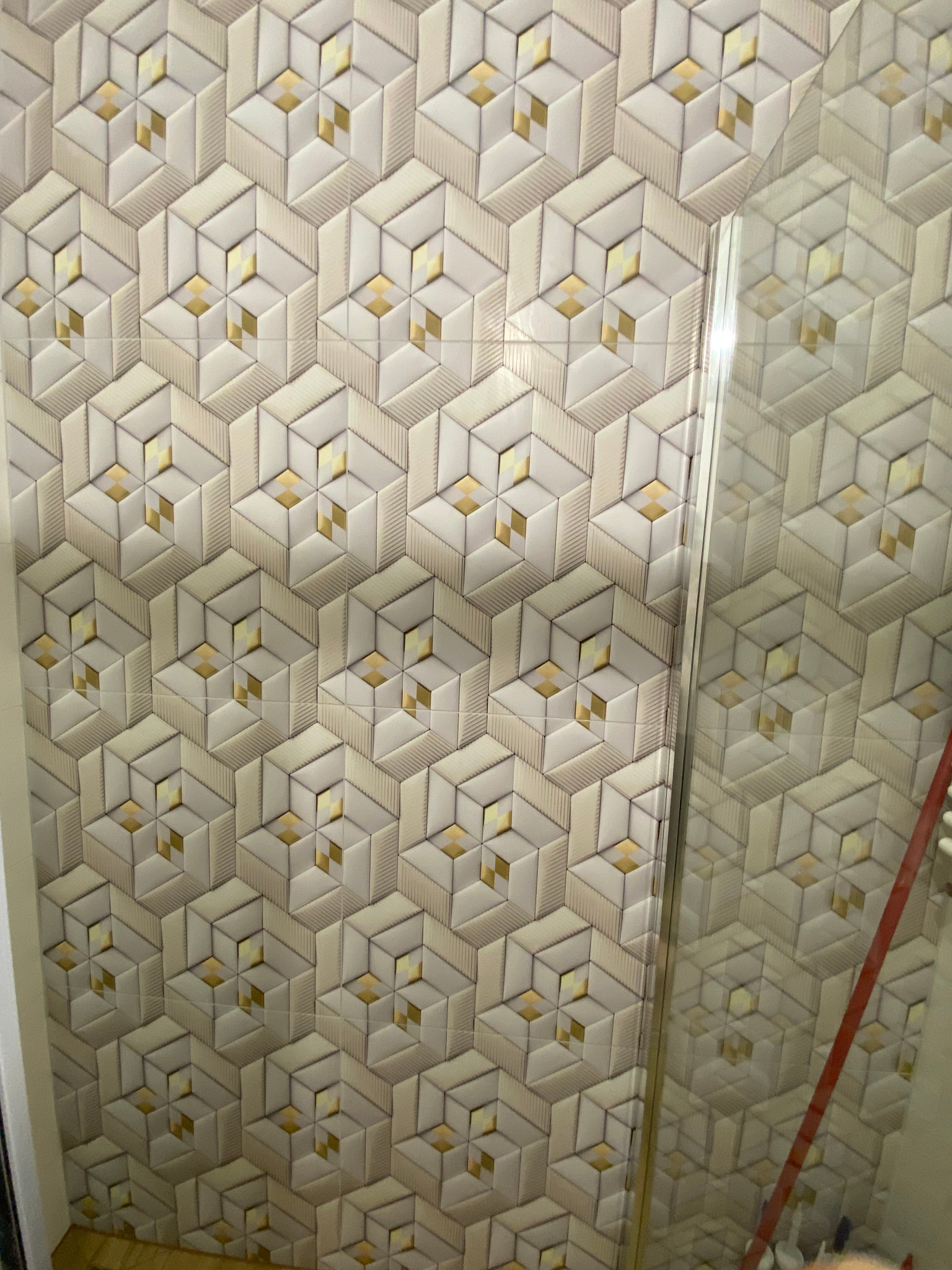 Ściana wyłożona trójwymiarowymi, heksagonalnymi płytkami ceramicznymi w odcieniach beżu i złota, odbicie w szklanej powierzchni.