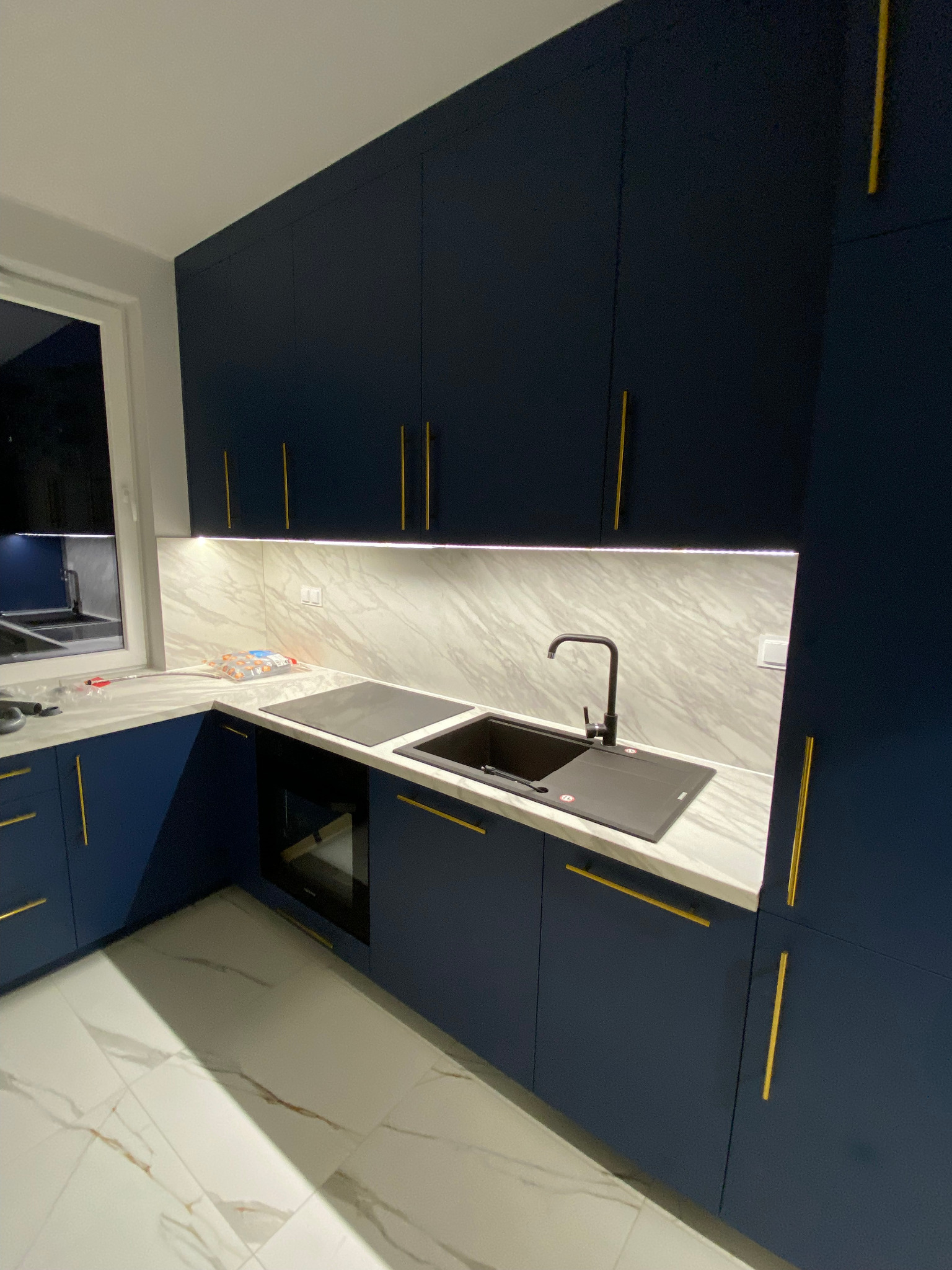 Nowoczesna, granatowa kuchnia z marmurowym blatem i złotymi uchwytami, czarny zlew i bateria, płyta indukcyjna, oświetlenie LED pod szafkami.