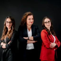 Trzy uśmiechnięte prawniczki w eleganckich marynarkach (czarnej, granatowej i czerwonej) stoją z założonymi rękami na tle jednolitego, czarnego tła.