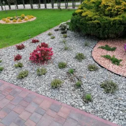 Ogród z różnorodnymi roślinami, krzewami i kwiatami, wysypany szarym i różowym żwirem, obok ścieżka z kostki brukowej.