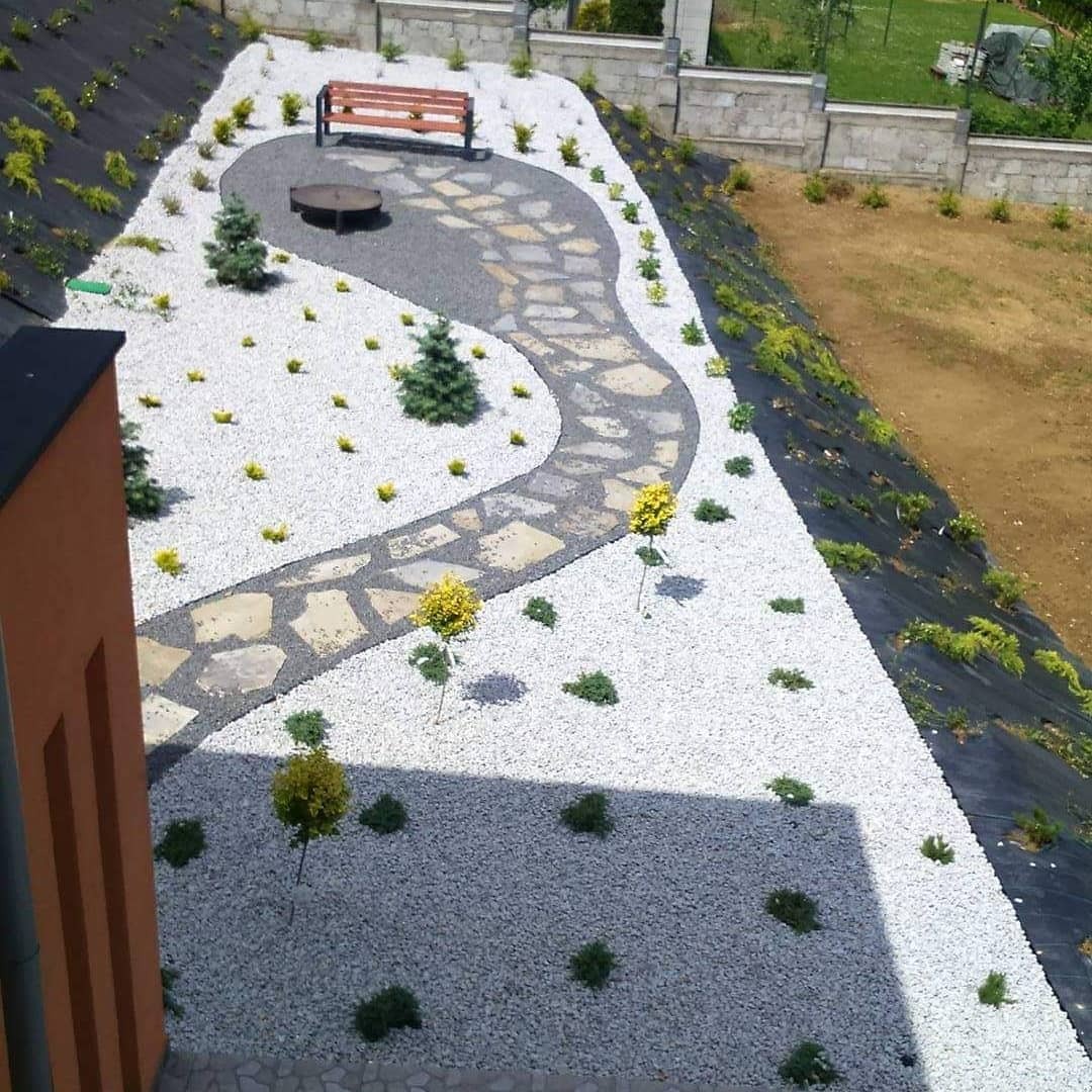 Ogród z krętą ścieżką z kamienia, białym żwirem, młodymi drzewkami i miejscem na ognisko, widok z góry.