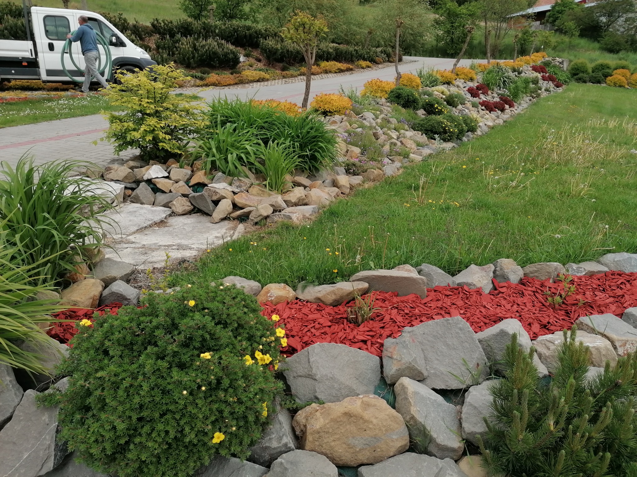 Ogród z kamieniami, roślinami i czerwoną korą, z pracownikiem i samochodem dostawczym w tle.