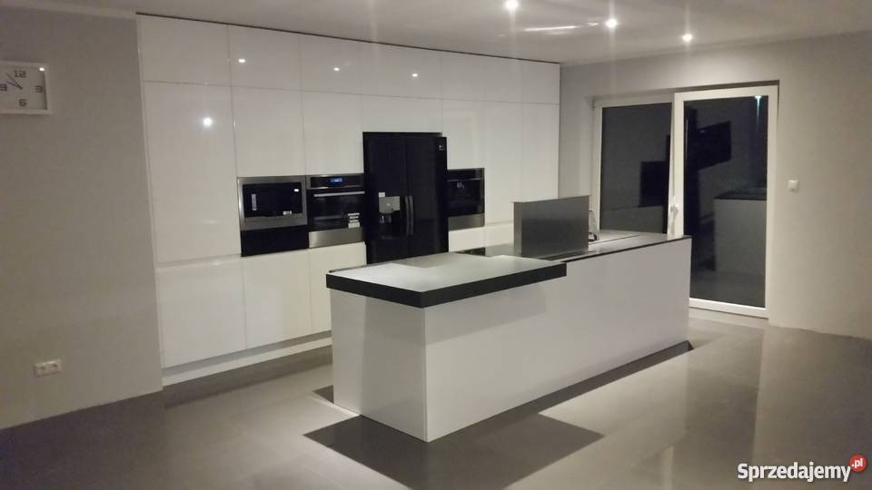 Nowoczesna kuchnia z białymi, lakierowanymi frontami, czarnym sprzętem AGD w zabudowie i wyspą kuchenną z czarnym blatem, widok na przesuwne drzwi balkonowe.