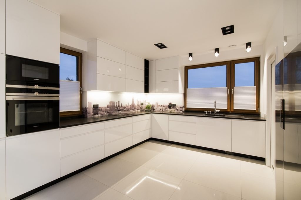 Nowoczesna, biała kuchnia z czarnym blatem i fototapetą z motywem panoramy miasta między szafkami górnymi i blatem, oświetlona punktowymi lampami sufitowymi i listwą LED pod szafkami.