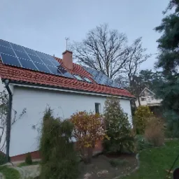 Biały dom z czerwoną dachówką i panelami słonecznymi na dachu, widok z poziomu ogrodu, pochmurny dzień.