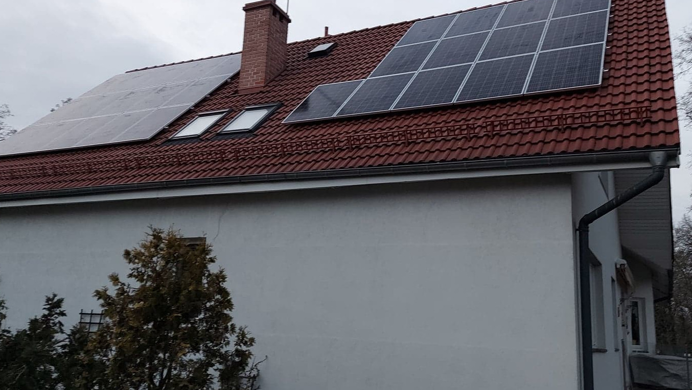 Czerwony dach z dachówki ceramicznej z zainstalowanymi panelami słonecznymi i oknami dachowymi, widok na komin z cegły i część elewacji budynku w pochmurny dzień.