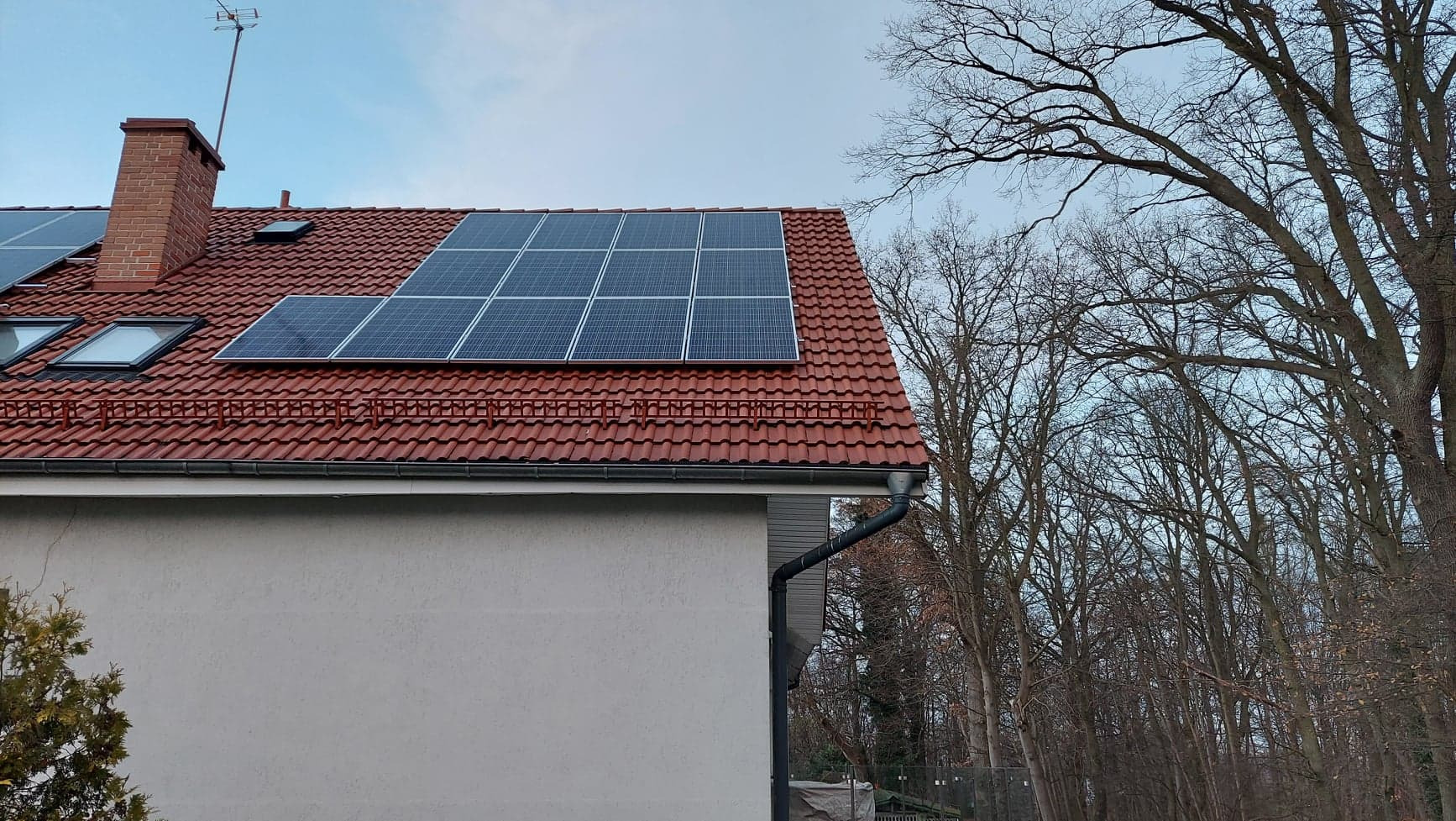 Panel słoneczny na dachu domu z czerwoną dachówką, widok z dołu na tle drzew bez liści.