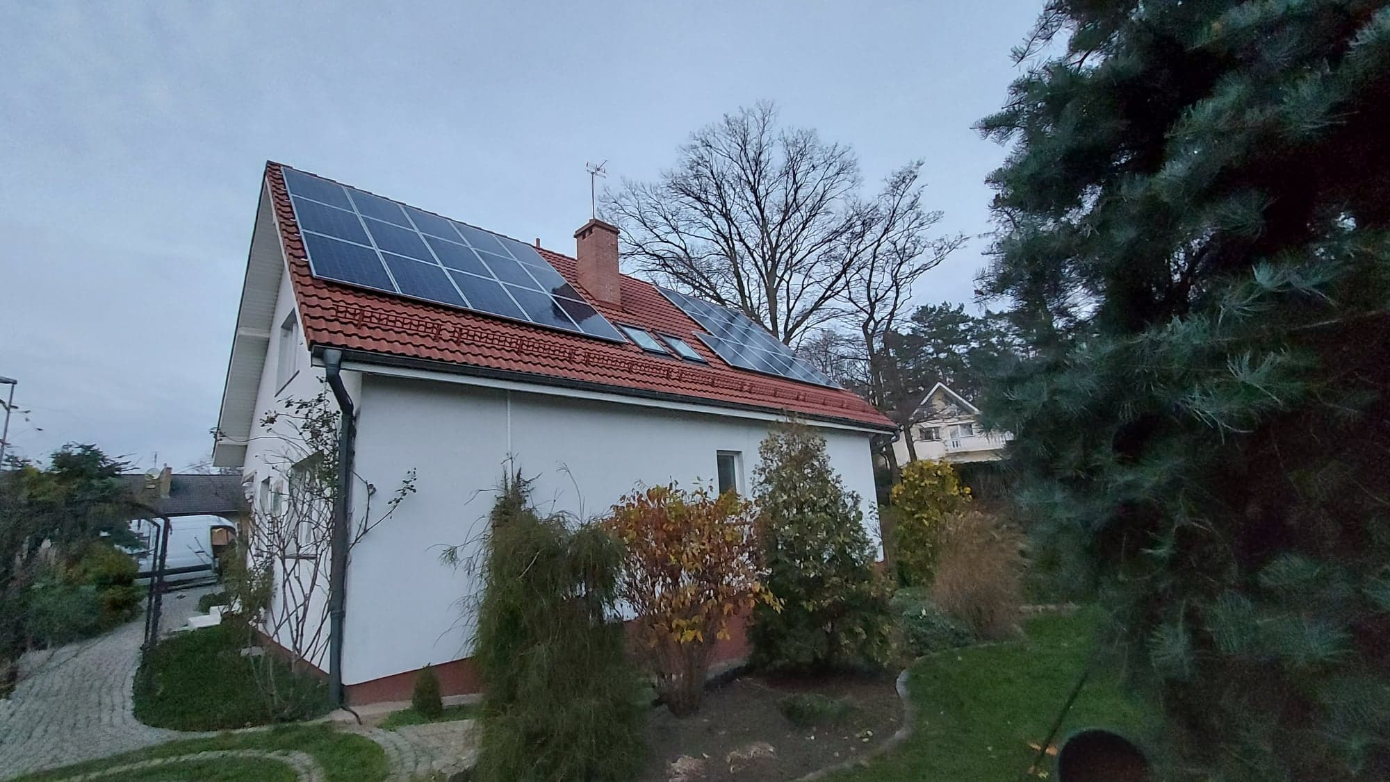 Biały dom z czerwoną dachówką i panelami słonecznymi na dachu, widok z poziomu ogrodu, pochmurny dzień.