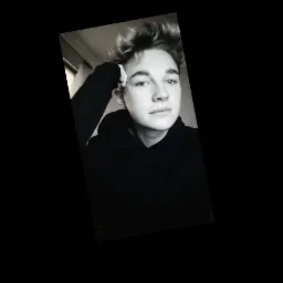 Portret młodego mężczyzny z blond włosami, ubranego w czarną bluzę z kapturem, opierającego głowę o rękę, z niebieskimi oczami i delikatnym uśmiechem.