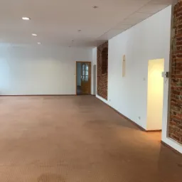 Puste pomieszczenie biurowe z białymi ścianami, ceglanymi elementami dekoracyjnymi i wykładziną dywanową w odcieniach brązu, widoczne oświetlenie sufitowe i drewniane drzwi w tle.