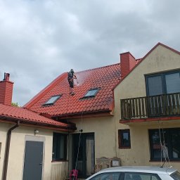 Hydrodynamiczny JAKUB SUROWANIEC - Mężczyzna w uprzęży myje czerwoną dachówkę domu z dwoma oknami dachowymi za pomocą myjki ciśnieniowej, widok z dołu na pochmurne niebo.