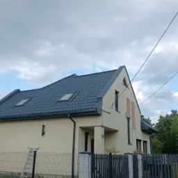 Dom jednorodzinny po renowacji z ciemnym dachem i jasną elewacją, widoczny z boku, z fragmentem rusztowania i drabiny. W tle żółty samochód dostawczy.