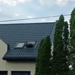 Ciemny dach z dachówką i otwartym oknem dachowym na tle jasnej elewacji budynku w trakcie budowy i zielonych drzew.