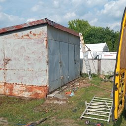 Hydrodynamiczny JAKUB SUROWANIEC - Malowanie metalowego garażu z widoczną rdzą przez osobę w masce, z otwartym żółtym vanem w tle, drabina oparta o ścianę, teren trawiasty.