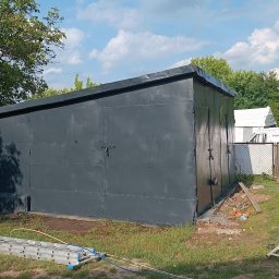 Hydrodynamiczny JAKUB SUROWANIEC - Malowanie metalowego garażu na szaro. Mężczyzna z wężem ogrodowym w tle, drabina i inne narzędzia na trawie. Widoczne ślady farby na ziemi i ścianie.