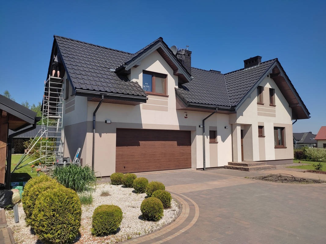 Dwukondygnacyjny dom jednorodzinny z szarą dachówką i elewacją w odcieniach beżu i brązu, z pracownikiem na aluminiowej drabinie przy dachu, na tle błękitnego nieba.