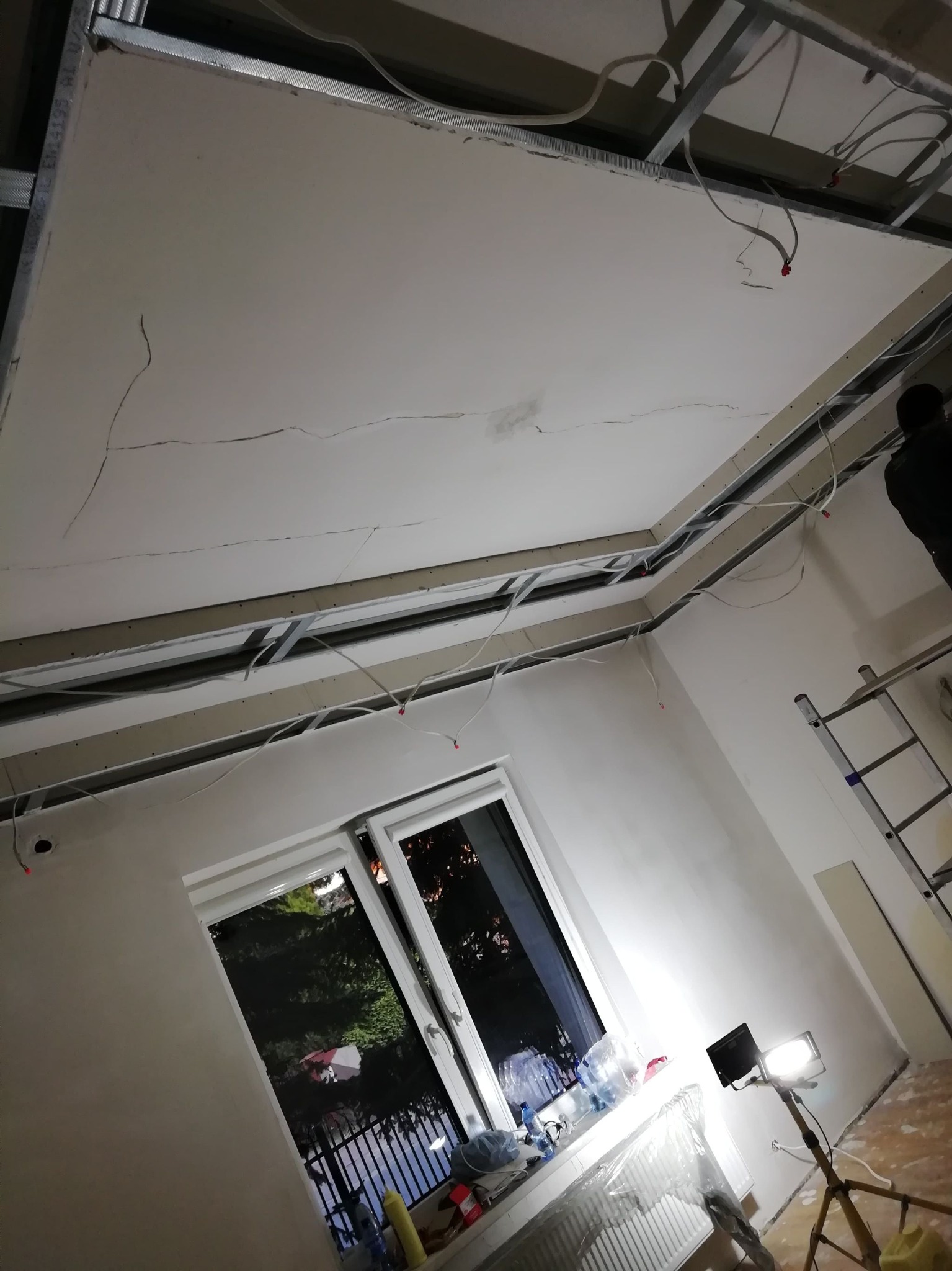 Montaż sufitu podwieszanego z płyt gipsowo-kartonowych, widoczne metalowe profile, instalacja elektryczna i pęknięcia na powierzchni płyty, okno z widokiem na zieleń.