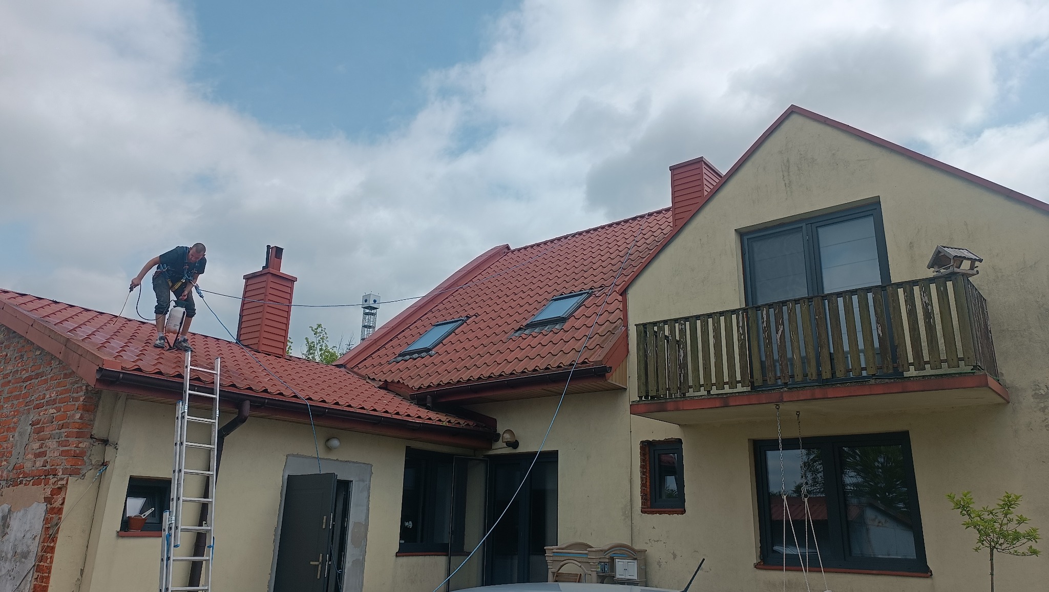 Pracownik w uprzęży, na drabinie, czyści czerwoną dachówkę domu z lukarnami. Widoczny budynek z balkonem i fragment elewacji z cegły. Czyszczenie dachu.