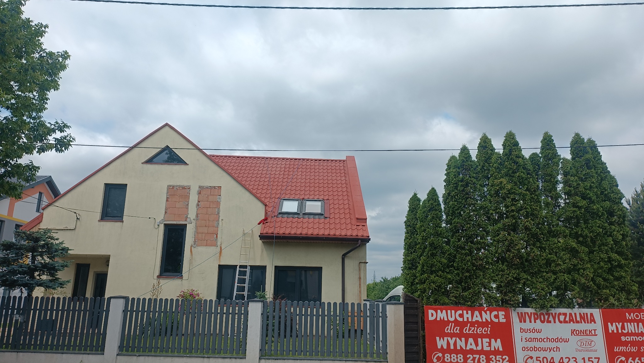 Dom jednorodzinny w trakcie prac elewacyjnych: fragmenty muru odsłonięte, drabina oparta o ścianę, czerwona dachówka. Widoczne reklamy na płocie.