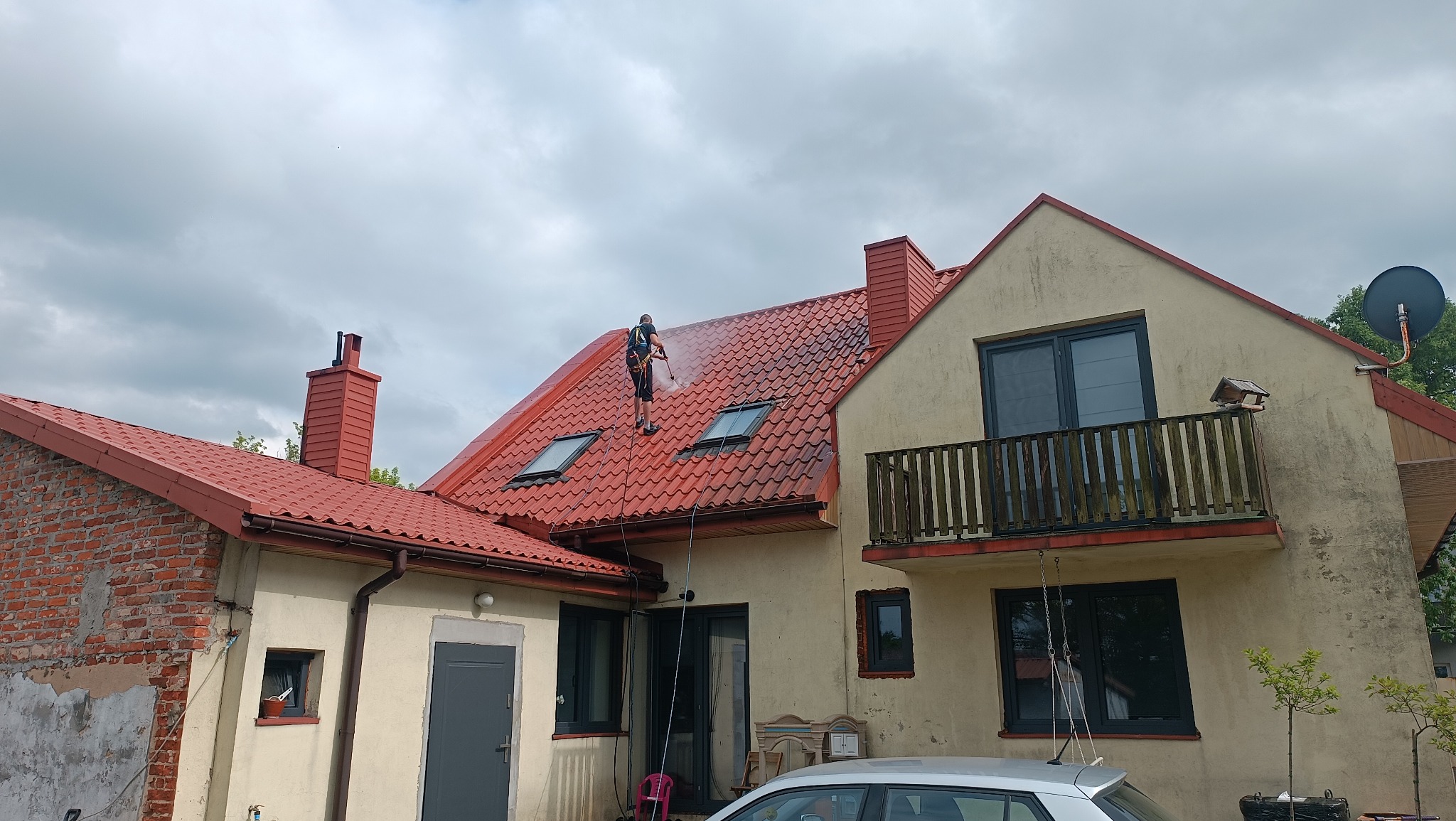 Mężczyzna w uprzęży myje czerwoną dachówkę domu z dwoma oknami dachowymi za pomocą myjki ciśnieniowej, widok z dołu na pochmurne niebo.