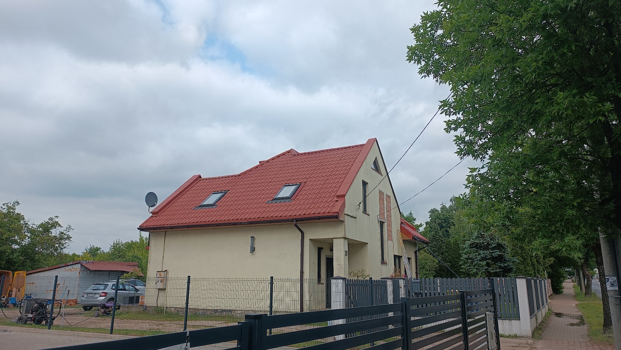 Jasny dom jednorodzinny z czerwoną dachówką i szarym ogrodzeniem. Widoczne okna dachowe, elewacja po renowacji i błękitne niebo z chmurami w tle.