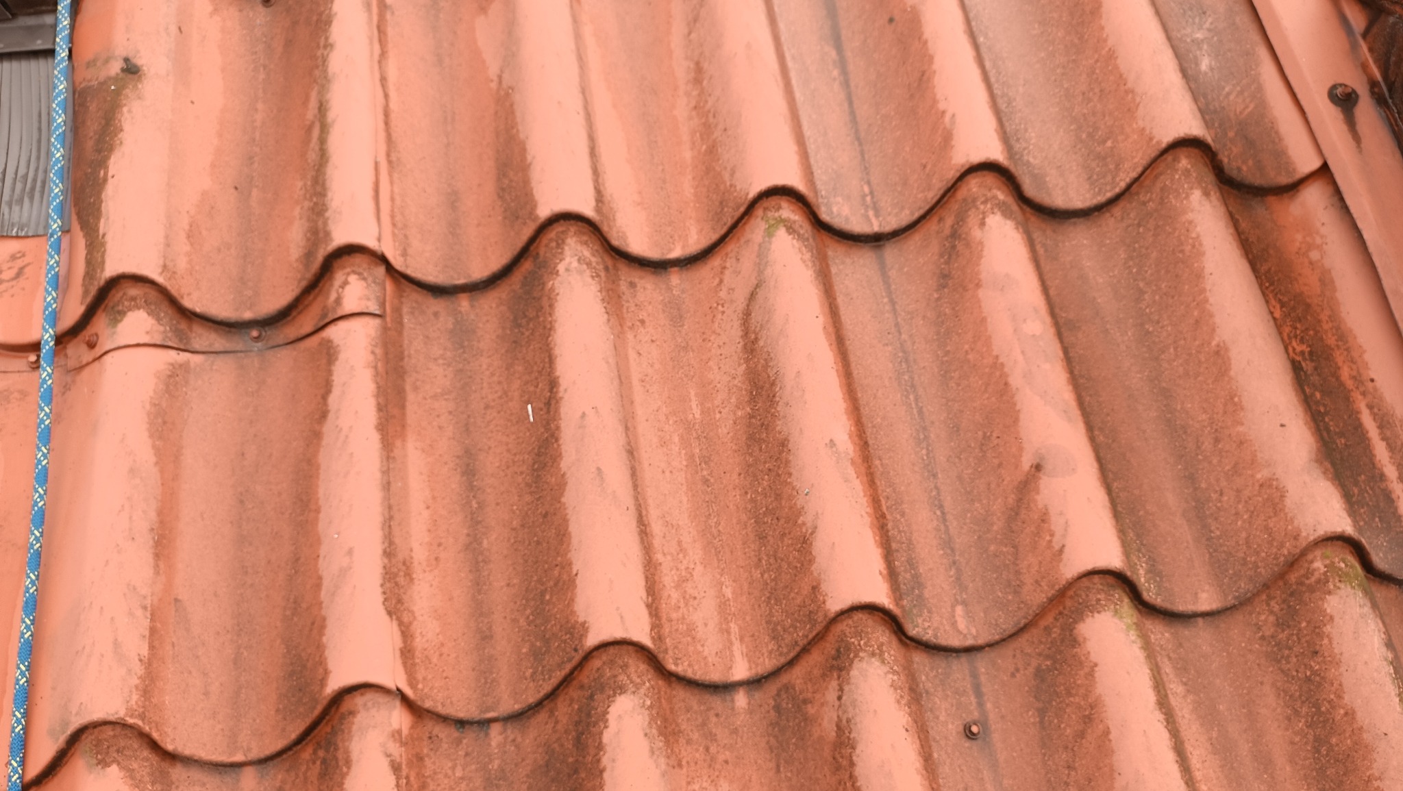 Czerwona dachówka ceramiczna z widocznymi śladami zabrudzeń i mchu. Na lewo widoczny fragment liny zabezpieczającej.