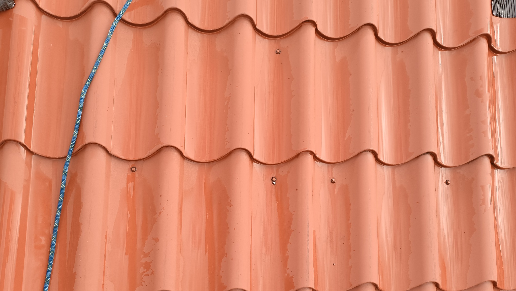 Błyszcząca, ceramiczna dachówka w odcieniach pomarańczy, z widoczną liną zabezpieczającą, sugerująca prace na wysokości. Detal mocowania dachówki w centralnej części kadru.