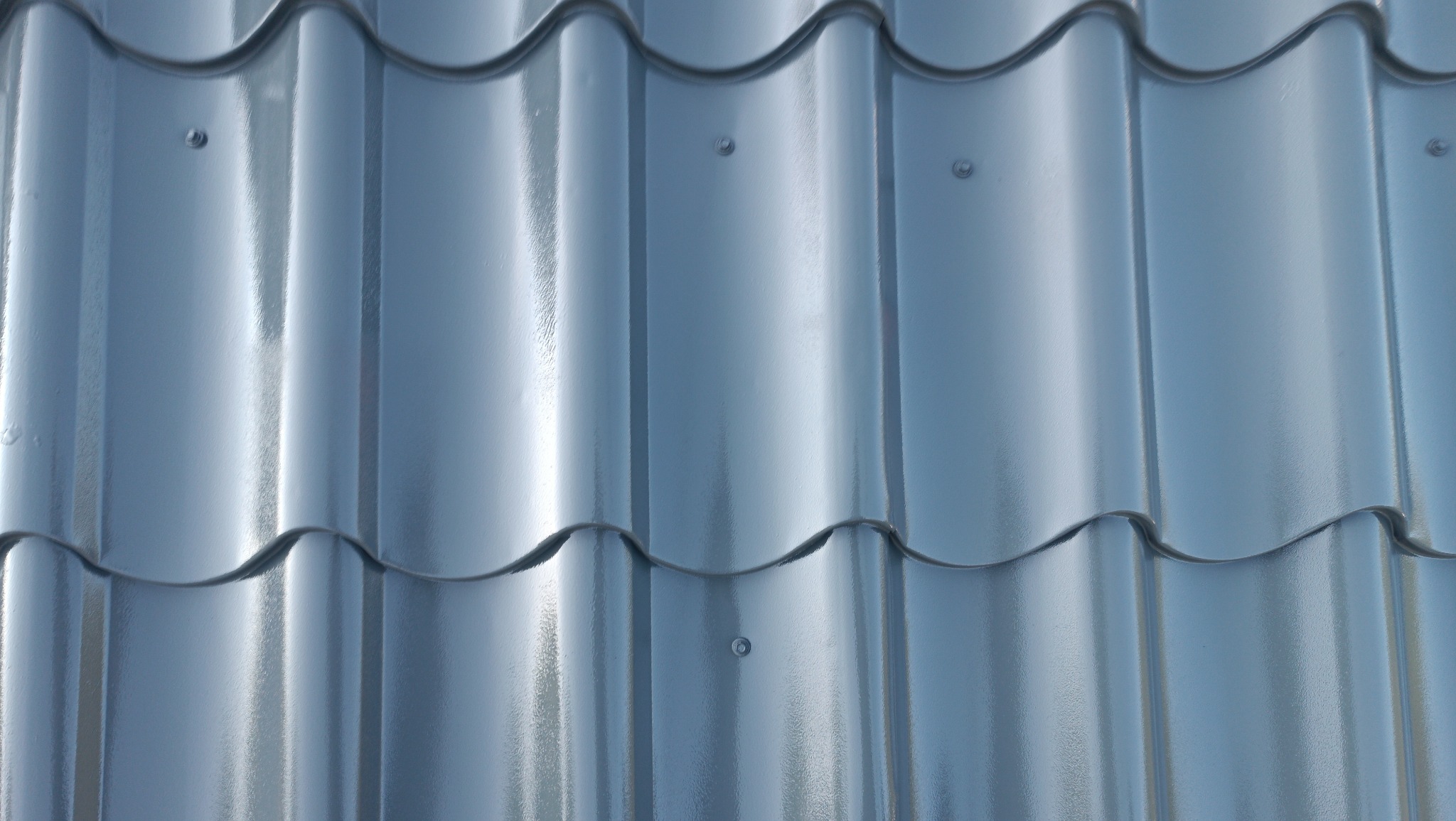 Błyszczące, szare dachówki metalowe, ułożone rzędami, z widocznymi punktami mocowania. Widok z bliska, ukazujący fakturę i refleksy świetlne.